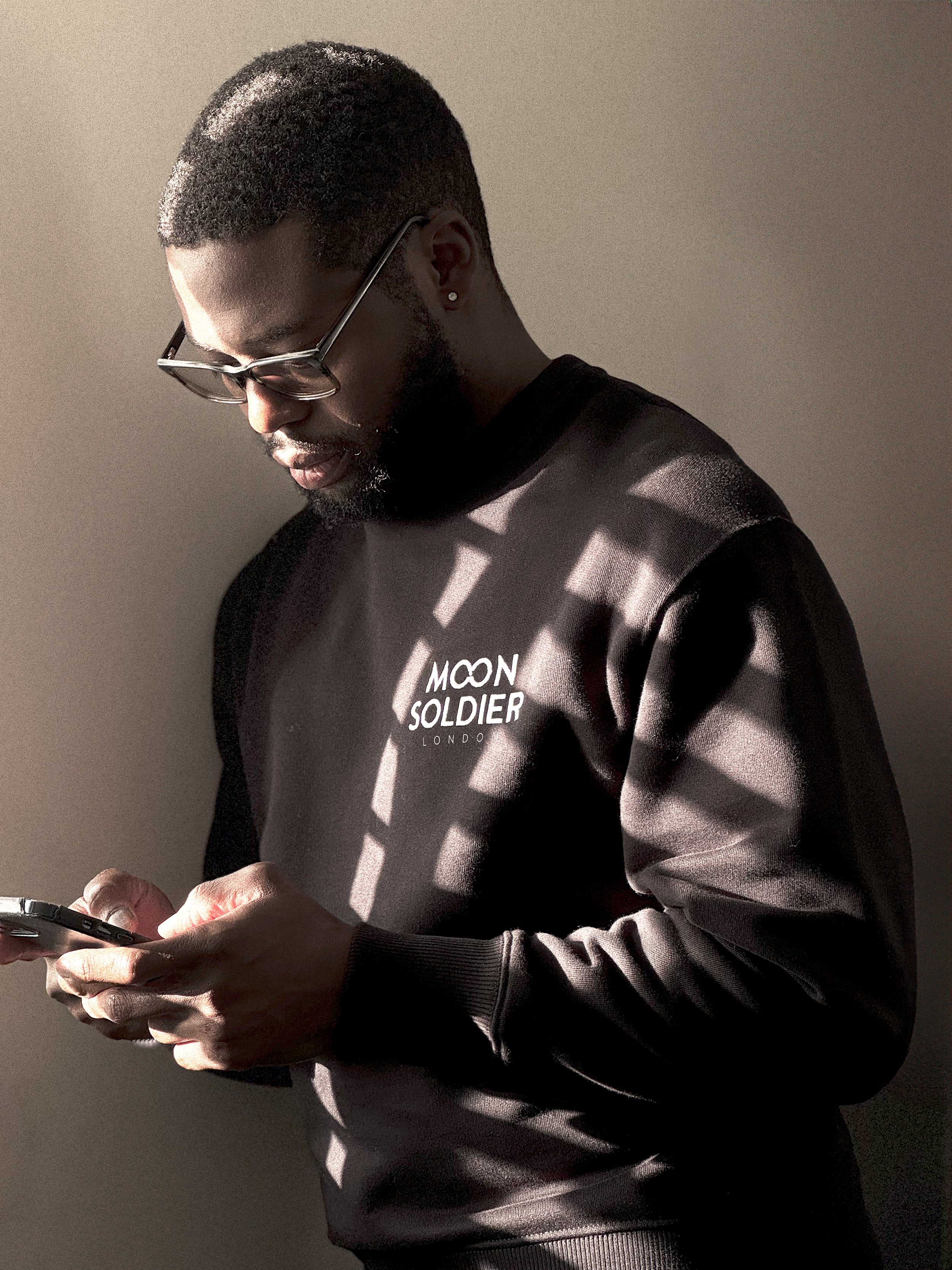 Header image showing a young African American male in glasses, focused on his phone in hand. He's wearing a black Moon Soldier sweatshirt with the logo on the left chest. The background is a warm grey wall.