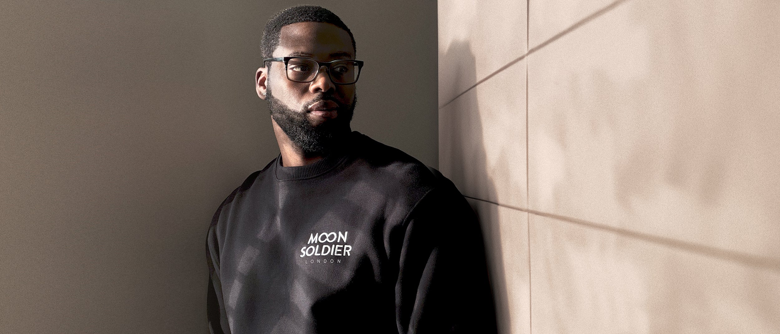 Header image featuring a young African American male wearing glasses and a black Moon Soldier sweatshirt, adorned with the Moon Soldier London logo on the left chest. He's positioned against a room corner, with one wall in warm grey and the other in muted soft pink panelling.